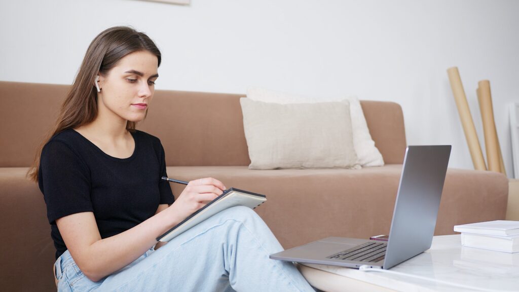 female student writng notes while looking at laptop placed on table