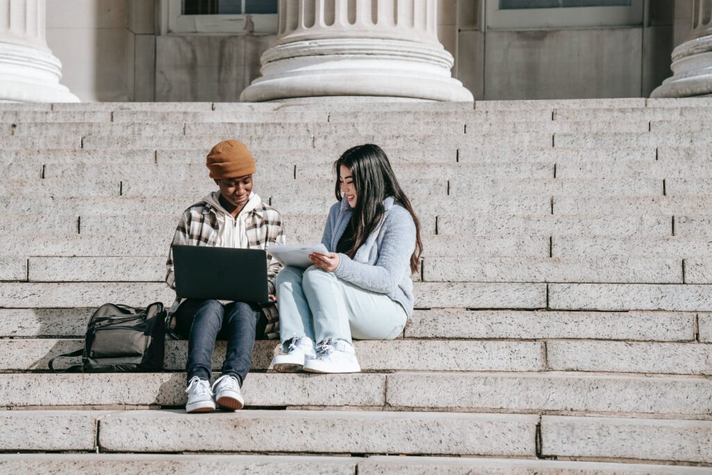 two femals sitting outside on a large staircase with laptop